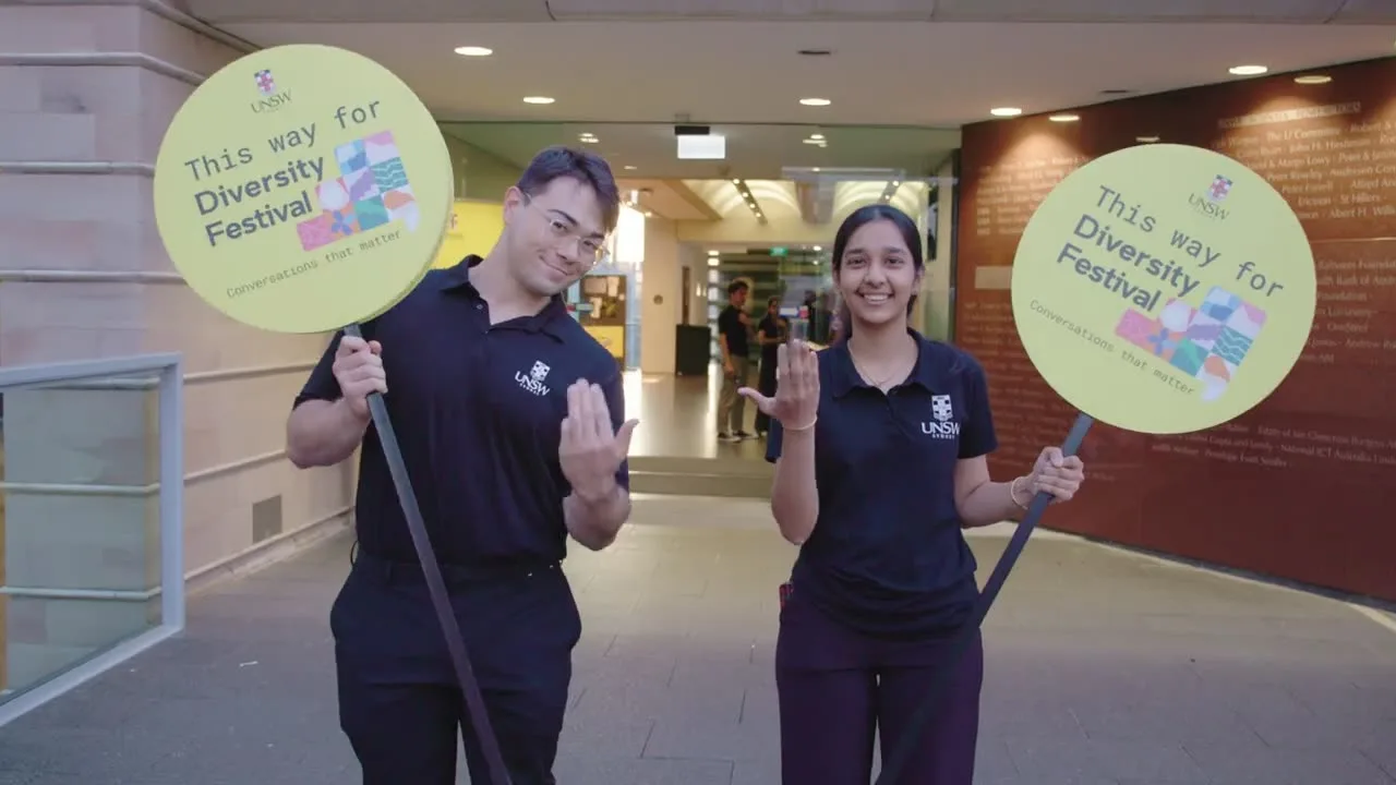 Two UNSW student volunteers in navy polos hold yellow 'This way for Diversity Festival' signs at a campus building entrance
